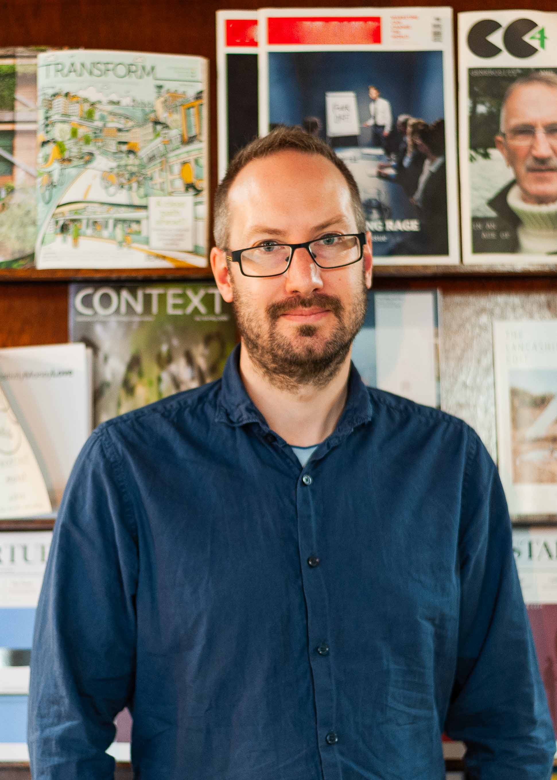 Team Alma: Ed Walker is standing in front of a bookcase of magazines, looking at the camera