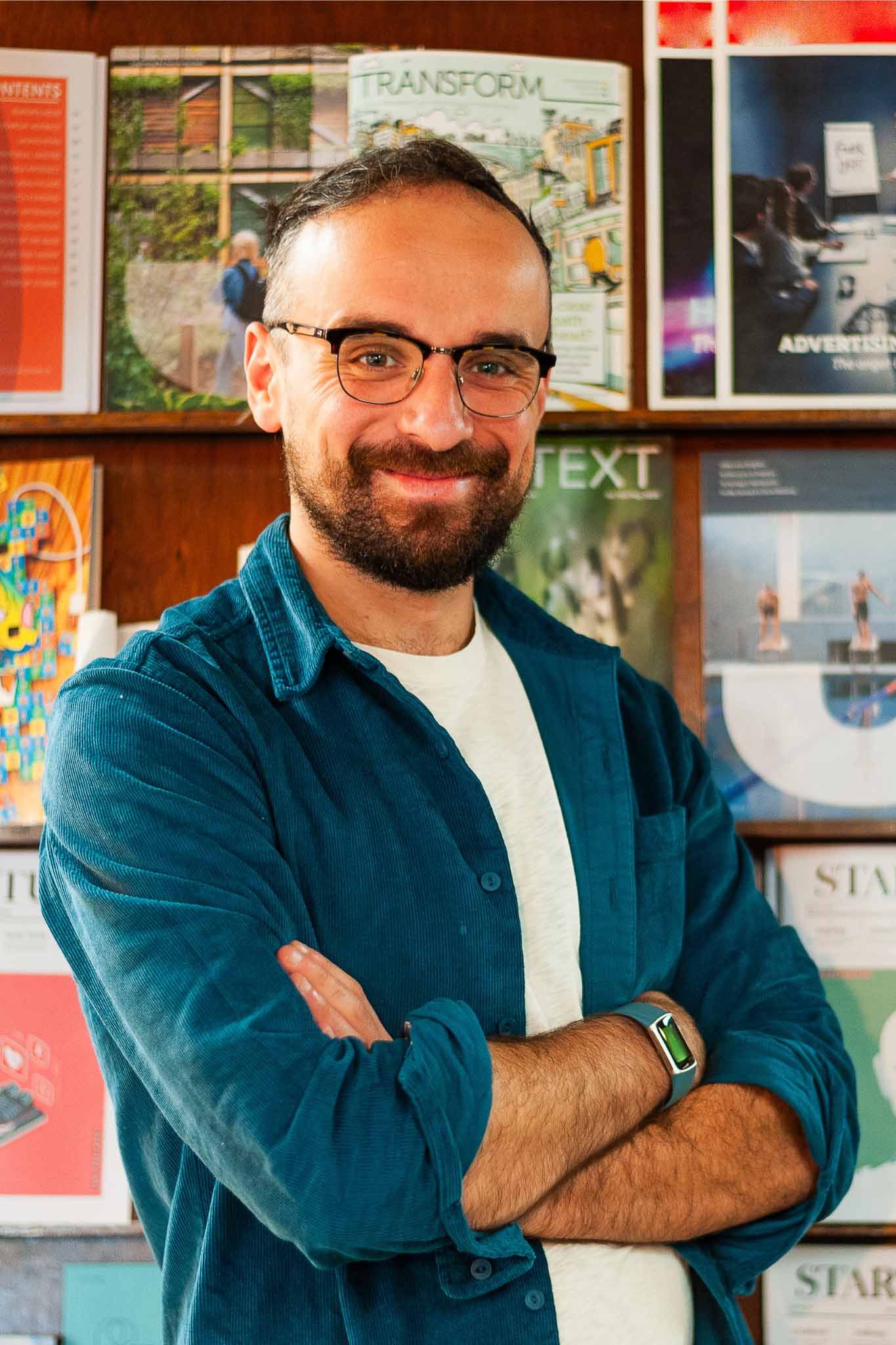 Team Alma: Luke Beardsworth is standing in front of a bookcase of magazines, with his arms folded, smiling at the camera