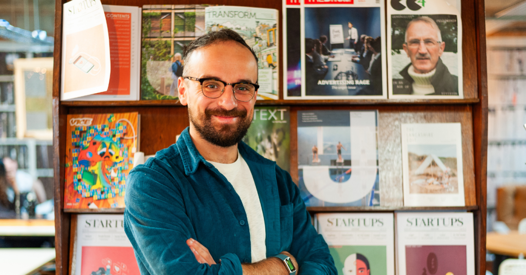 Luke smiling at the camera, arms folded. Background is a bookcase with magazines displayed.