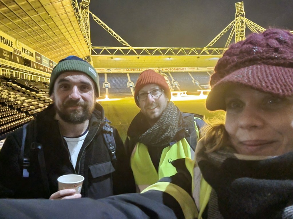 The photo is a selfie with Luke Beardsworth, Ed Walker and Shirah Bamber looking at camera, in winter hats. The background shows the PNE football stadium at night behind them.