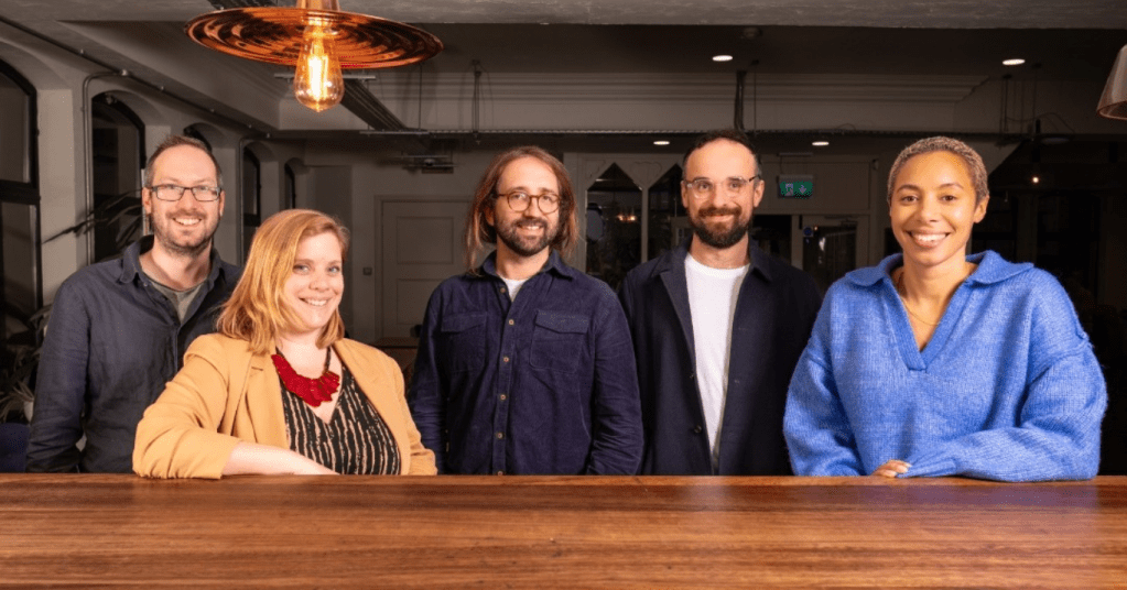 In the photo Ed Walker, Shirah Bamber, Dimi Reider, Luke Beardsworth and Natalie Morris are standing in front of a wooden countertop, smiling at the camera.