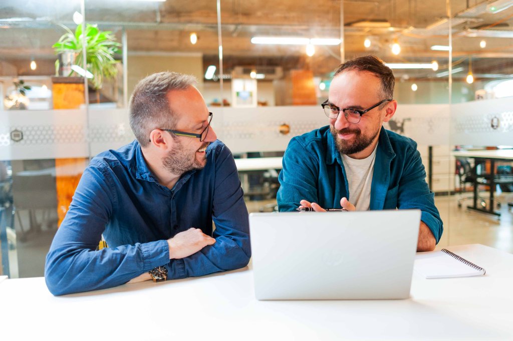 Ed and Luke are in an office space, sitting at a desk with a laptop in front of them. They are wearing blue shirts and smiling at each other.