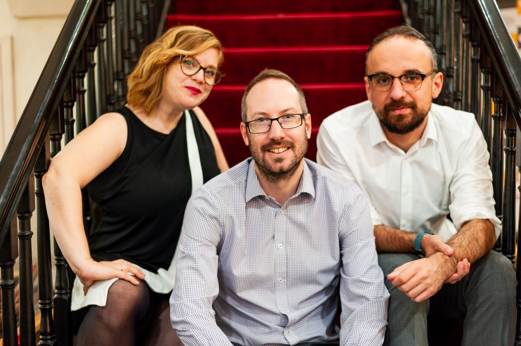 Team Alma (Shirah Bamber, Ed Walker, and Luke Beardsworth) are sitting on wooden stairs with dark red carpet.

They are all looking at the camera.