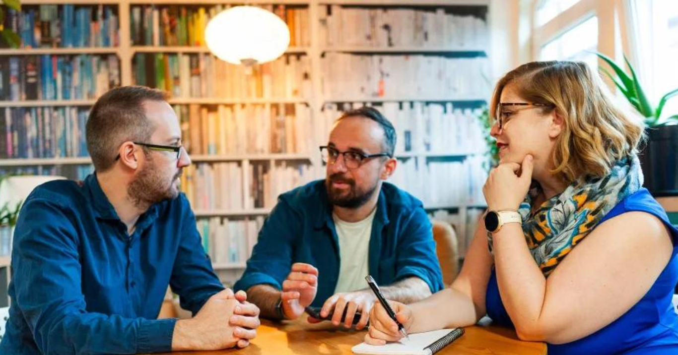 Ed Walker, Luke Beardsworth and Shirah Bamber are sitting at a table, interacting with each other.