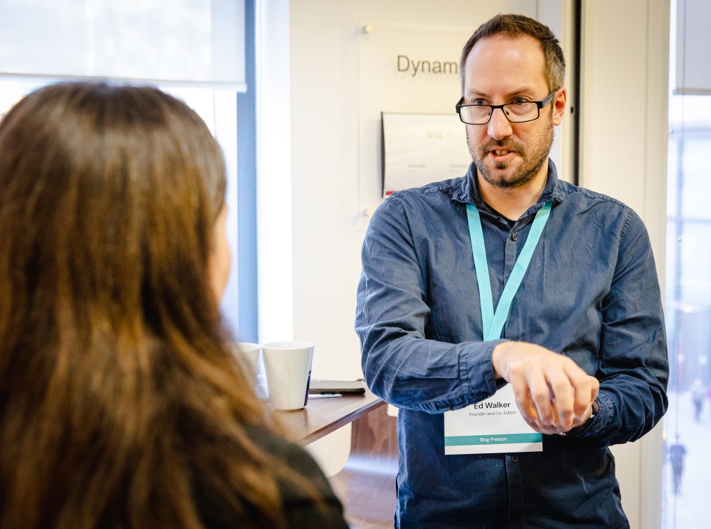 Ed is standing, facing the camera, in a conversation with someone whose back is to the camera. He is wearing a blue shirt and a name tag, his hands are mid-action.